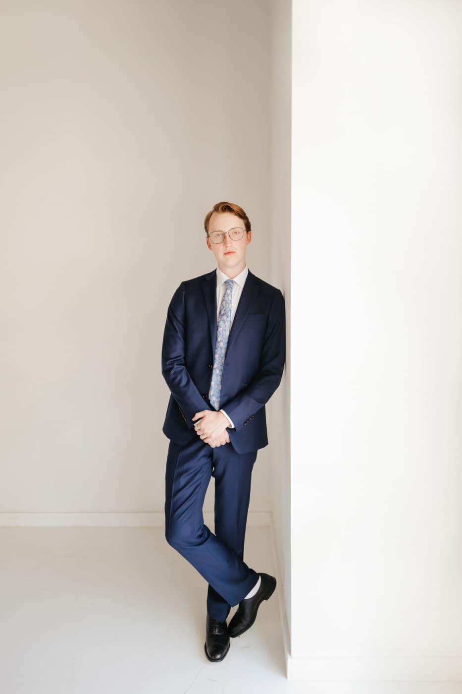 A young man in a navy suit leaning against a white wall