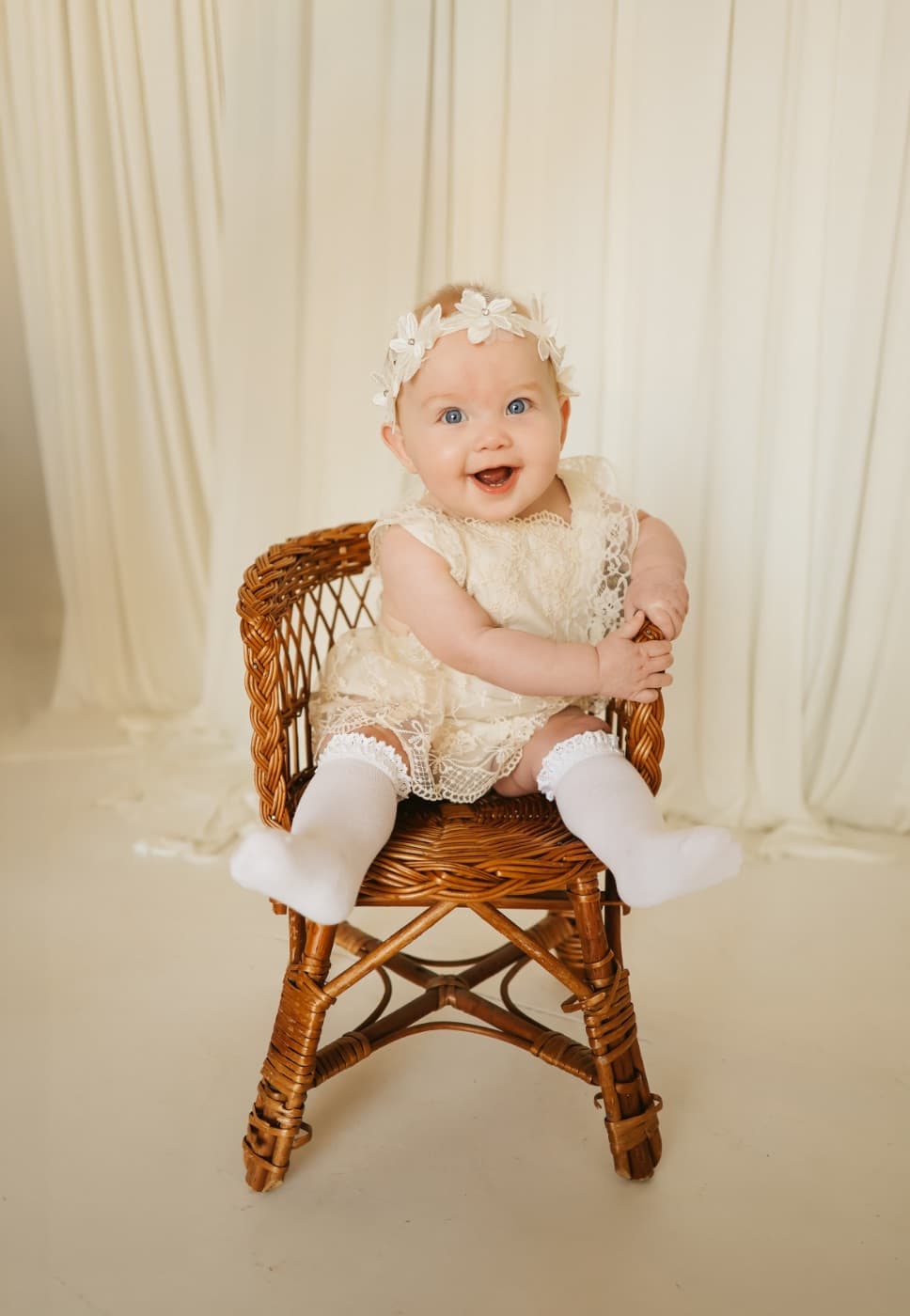 A baby in a lace dress on a wicker chair in front of cream drapes